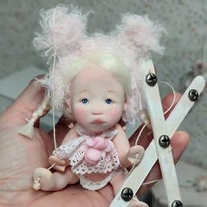 Op de afbeelding: Een handgemaakte pop met een roze, pluizige hoed en een witte kanten outfit. De pop heeft blauwe ogen, roze wangen en blond haar. Ze zit op een miniatuur schommel gemaakt van wit geschilderd hout en bij elkaar gehouden met schroeven.