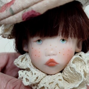 Puede incluir: Cabeza de mu&ntilde;eca con cabello casta&ntilde;o, ojos azules y pecas. La mu&ntilde;eca lleva un sombrero beige con un estampado floral y un cuello de encaje color crema. Los labios de la mu&ntilde;eca son de color coral.