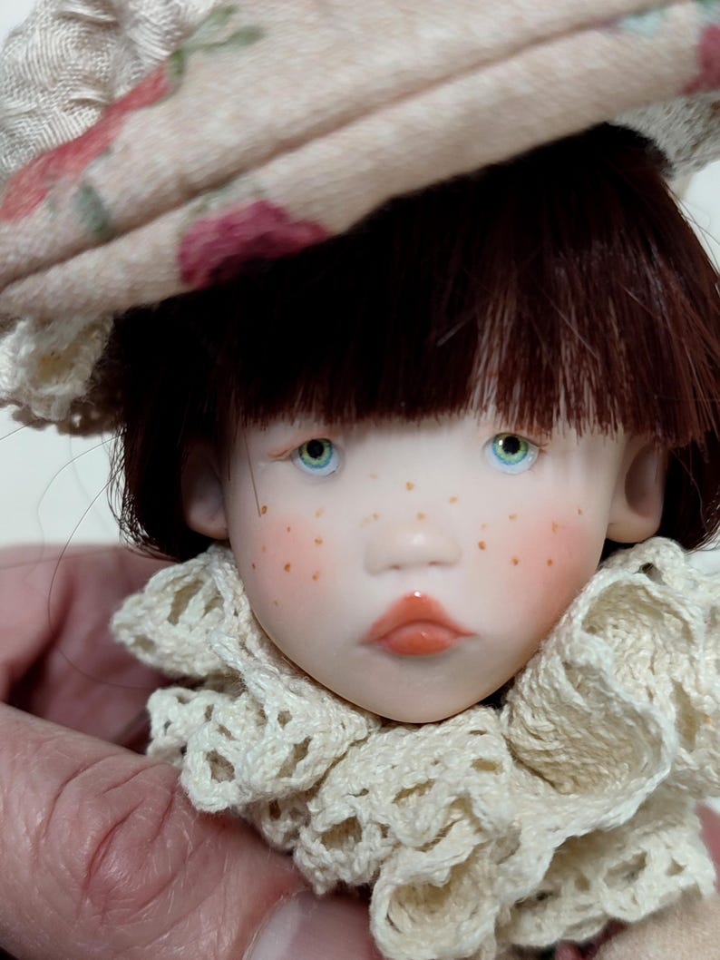 Puede incluir: Cabeza de mu&ntilde;eca con cabello casta&ntilde;o, ojos azules y pecas. La mu&ntilde;eca lleva un sombrero beige con un estampado floral y un cuello de encaje color crema. Los labios de la mu&ntilde;eca son de color coral.