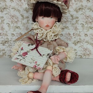 Puede incluir: Una mu&ntilde;eca artesanal con un rostro detallado, que lleva un gorro y un cuello con volantes. La mu&ntilde;eca sostiene un peque&ntilde;o libro con un dise&ntilde;o floral. El atuendo de la mu&ntilde;eca es una mezcla de colores beige y crema, con detalles en rojo en las sandalias.