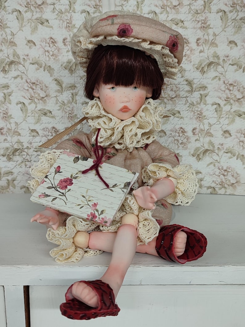 Puede incluir: Una mu&ntilde;eca artesanal con un rostro detallado, que lleva un gorro y un cuello con volantes. La mu&ntilde;eca sostiene un peque&ntilde;o libro con un dise&ntilde;o floral. El atuendo de la mu&ntilde;eca es una mezcla de colores beige y crema, con detalles en rojo en las sandalias.