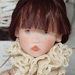 Puede incluir: Una mu&ntilde;eca artesanal con rostro de porcelana, ojos celeste claro y cabello casta&ntilde;o rojizo. La mu&ntilde;eca tiene pecas y lleva un cuello de ganchillo color crema y un vestido floral rosa. Los labios de la mu&ntilde;eca est&aacute;n pintados de color melocot&oacute;n claro.