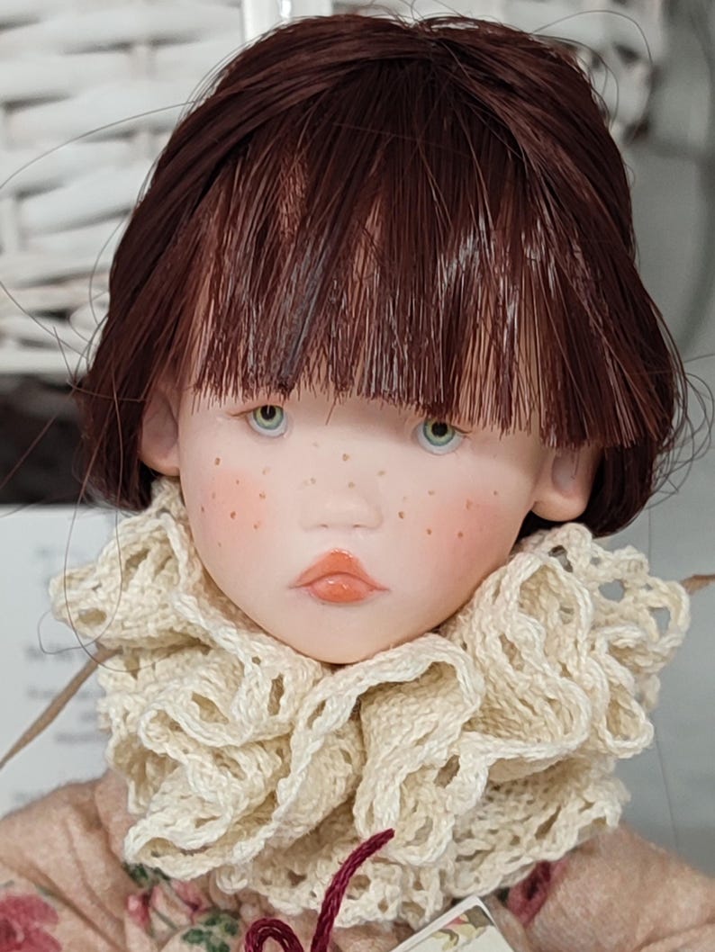 Puede incluir: Una mu&ntilde;eca artesanal con rostro de porcelana, ojos celeste claro y cabello casta&ntilde;o rojizo. La mu&ntilde;eca tiene pecas y lleva un cuello de ganchillo color crema y un vestido floral rosa. Los labios de la mu&ntilde;eca est&aacute;n pintados de color melocot&oacute;n claro.