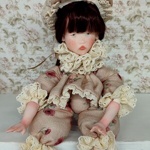 Puede incluir: Mu&ntilde;eca hecha a mano con un atuendo beige adornado con patrones florales rojos. La mu&ntilde;eca tiene un sombrero a juego y un delicado ribete de encaje con volantes en el cuello, las mangas y los pu&ntilde;os de los pantalones. La mu&ntilde;eca tiene cabello casta&ntilde;o oscuro y ojos azules.