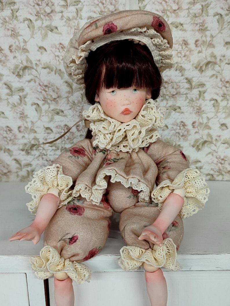 Puede incluir: Mu&ntilde;eca hecha a mano con un atuendo beige adornado con patrones florales rojos. La mu&ntilde;eca tiene un sombrero a juego y un delicado ribete de encaje con volantes en el cuello, las mangas y los pu&ntilde;os de los pantalones. La mu&ntilde;eca tiene cabello casta&ntilde;o oscuro y ojos azules.