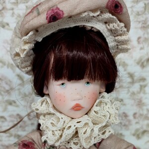 Puede incluir: Mu&ntilde;eca de piel clara, pecas y ojos azules, vestida con un vestido beige con estampado floral. La mu&ntilde;eca tiene cabello casta&ntilde;o rojizo y un sombrero beige con detalles florales rosas. Un cuello de encaje color crema enmarca la cara de la mu&ntilde;eca.