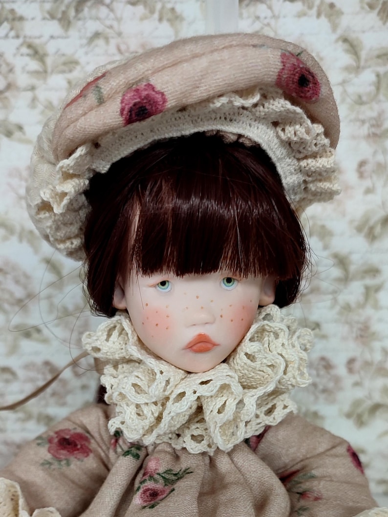 Puede incluir: Mu&ntilde;eca de piel clara, pecas y ojos azules, vestida con un vestido beige con estampado floral. La mu&ntilde;eca tiene cabello casta&ntilde;o rojizo y un sombrero beige con detalles florales rosas. Un cuello de encaje color crema enmarca la cara de la mu&ntilde;eca.