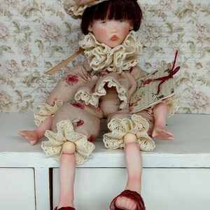 Puede incluir: Mu&ntilde;eca hecha a mano con rostro pintado, vestida con un atuendo beige con patrones de rosas rojas y adornos de encaje crema. La mu&ntilde;eca tiene un sombrero a juego y sandalias rojas. Sostiene un peque&ntilde;o libro con una cinta roja.