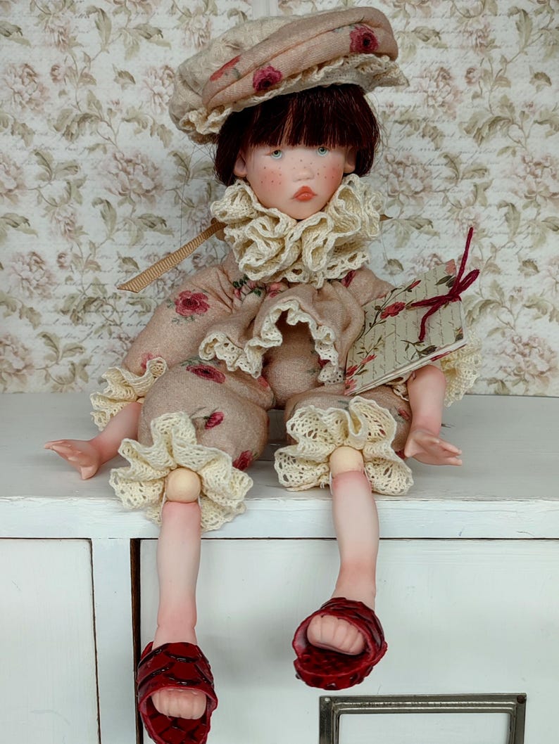 Puede incluir: Mu&ntilde;eca hecha a mano con rostro pintado, vestida con un atuendo beige con patrones de rosas rojas y adornos de encaje crema. La mu&ntilde;eca tiene un sombrero a juego y sandalias rojas. Sostiene un peque&ntilde;o libro con una cinta roja.