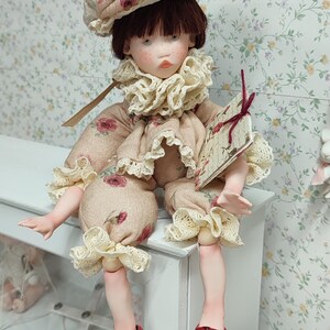 Puede incluir: Una mu&ntilde;eca hecha a mano con est&eacute;tica vintage. La mu&ntilde;eca viste un conjunto beige con estampado floral, un cuello con volantes y un sombrero a juego. La mu&ntilde;eca tiene sandalias rojas y sostiene un peque&ntilde;o libro. La mu&ntilde;eca est&aacute; sentada sobre una superficie blanca.