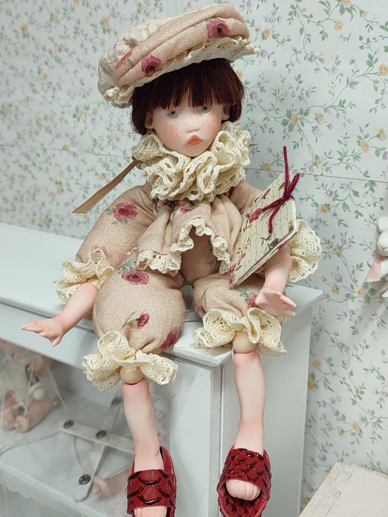 Puede incluir: Una mu&ntilde;eca hecha a mano con est&eacute;tica vintage. La mu&ntilde;eca viste un conjunto beige con estampado floral, un cuello con volantes y un sombrero a juego. La mu&ntilde;eca tiene sandalias rojas y sostiene un peque&ntilde;o libro. La mu&ntilde;eca est&aacute; sentada sobre una superficie blanca.