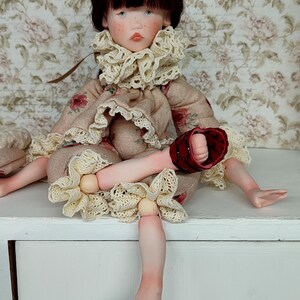 Puede incluir: Mu&ntilde;eca hecha a mano con rostro pintado, cabello oscuro y ojos azules. La mu&ntilde;eca viste un vestido floral con cuello con volantes y sandalias rojas. La mu&ntilde;eca est&aacute; sentada sobre una superficie blanca.