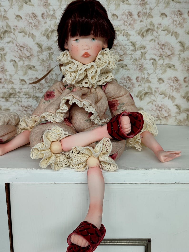 Puede incluir: Mu&ntilde;eca hecha a mano con rostro pintado, cabello oscuro y ojos azules. La mu&ntilde;eca viste un vestido floral con cuello con volantes y sandalias rojas. La mu&ntilde;eca est&aacute; sentada sobre una superficie blanca.