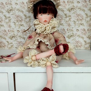 Puede incluir: Mu&ntilde;eca hecha a mano con un vestido beige y sombrero a juego, adornada con estampados florales y ribetes de encaje. La mu&ntilde;eca tiene cabello oscuro, ojos azules y sandalias rojas. La mu&ntilde;eca est&aacute; sentada sobre una superficie blanca.