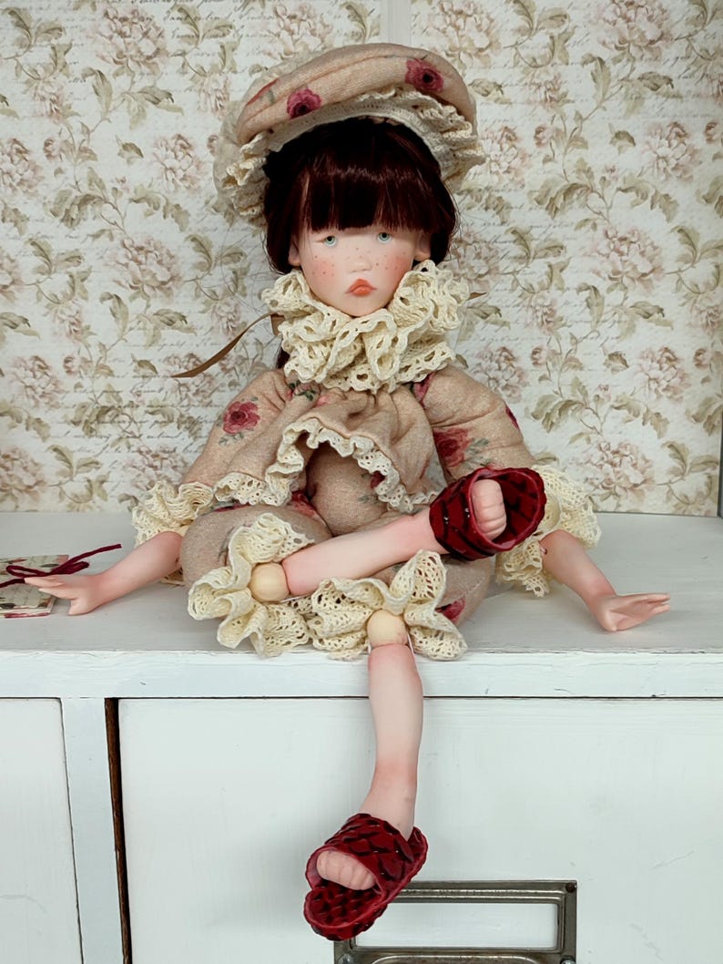 Puede incluir: Mu&ntilde;eca hecha a mano con un vestido beige y sombrero a juego, adornada con estampados florales y ribetes de encaje. La mu&ntilde;eca tiene cabello oscuro, ojos azules y sandalias rojas. La mu&ntilde;eca est&aacute; sentada sobre una superficie blanca.