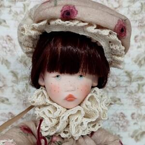 Puede incluir: Una mu&ntilde;eca hecha a mano con tez p&aacute;lida, pecas y ojos azules. La mu&ntilde;eca lleva un gorro beige con detalles florales rosas y un ribete de encaje color crema. Un vestido a juego y un cuello con volantes completan el look vintage.