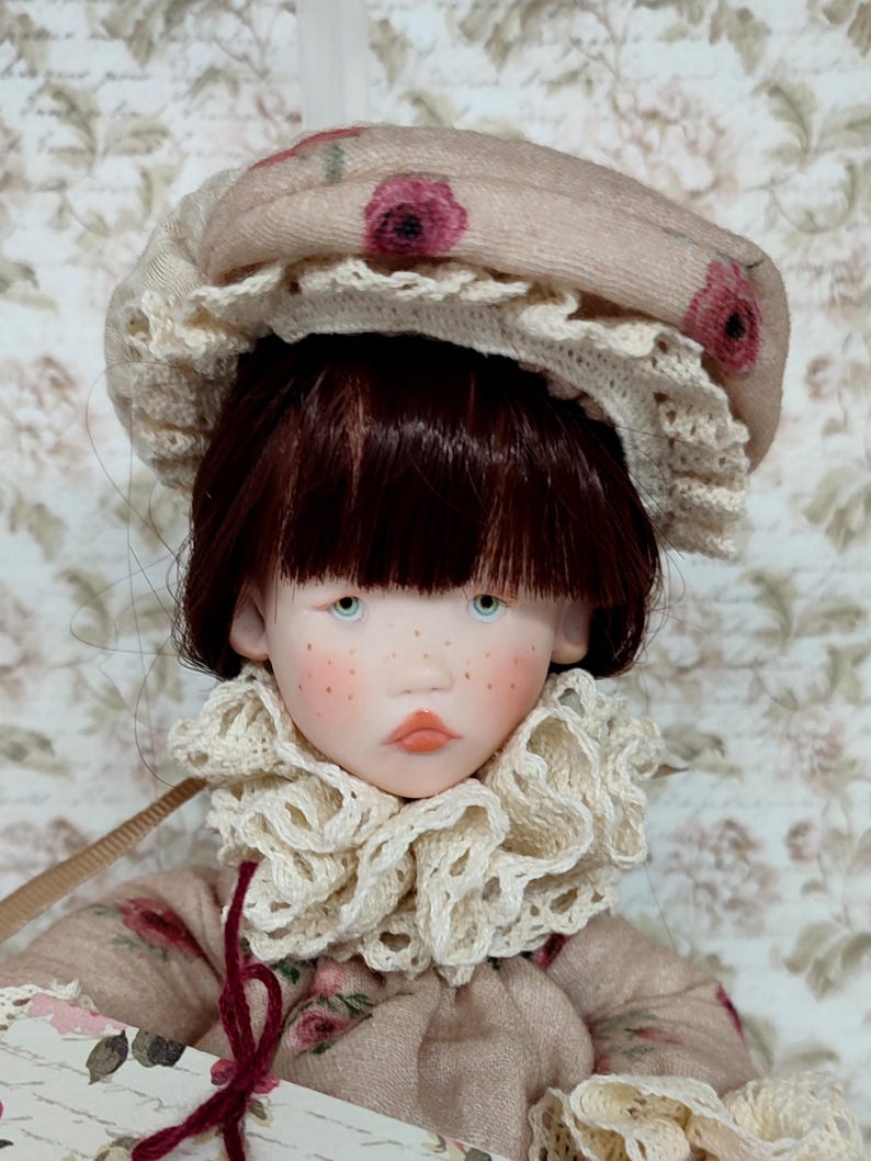Puede incluir: Una mu&ntilde;eca hecha a mano con tez p&aacute;lida, pecas y ojos azules. La mu&ntilde;eca lleva un gorro beige con detalles florales rosas y un ribete de encaje color crema. Un vestido a juego y un cuello con volantes completan el look vintage.