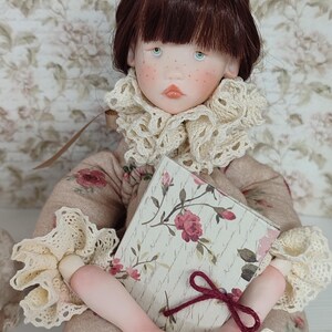 Puede incluir: Mu&ntilde;eca hecha a mano con cabello casta&ntilde;o, ojos azules y pecas, vestida con un atuendo beige con estampado floral con cuello y pu&ntilde;os de encaje color crema. La mu&ntilde;eca sostiene un libro con un dise&ntilde;o floral y una cinta burdeos.