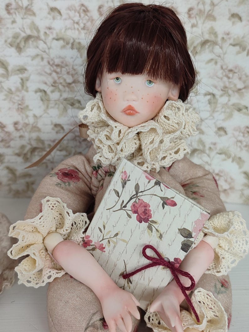 Puede incluir: Mu&ntilde;eca hecha a mano con cabello casta&ntilde;o, ojos azules y pecas, vestida con un atuendo beige con estampado floral con cuello y pu&ntilde;os de encaje color crema. La mu&ntilde;eca sostiene un libro con un dise&ntilde;o floral y una cinta burdeos.
