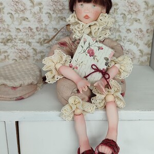 Puede incluir: Una mu&ntilde;eca sentada con cabello casta&ntilde;o y pecas, vestida con un atuendo con estampado floral con adornos de encaje color crema. La mu&ntilde;eca sostiene un libro con una cubierta floral y usa sandalias rojas. Un sombrero a juego est&aacute; a la izquierda.