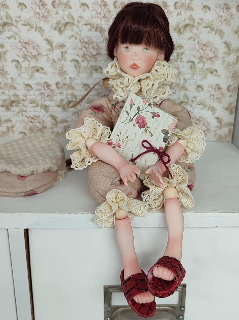 Puede incluir: Una mu&ntilde;eca sentada con cabello casta&ntilde;o y pecas, vestida con un atuendo con estampado floral con adornos de encaje color crema. La mu&ntilde;eca sostiene un libro con una cubierta floral y usa sandalias rojas. Un sombrero a juego est&aacute; a la izquierda.