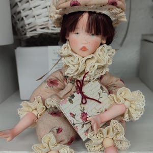 Puede incluir: Una mu&ntilde;eca hecha a mano con la cara pintada, vestida con un vestido con estampado floral, un sombrero a juego y un cuello y pu&ntilde;os con volantes. La mu&ntilde;eca sostiene un peque&ntilde;o libro con un dise&ntilde;o floral. La mu&ntilde;eca est&aacute; sentada sobre una superficie blanca.