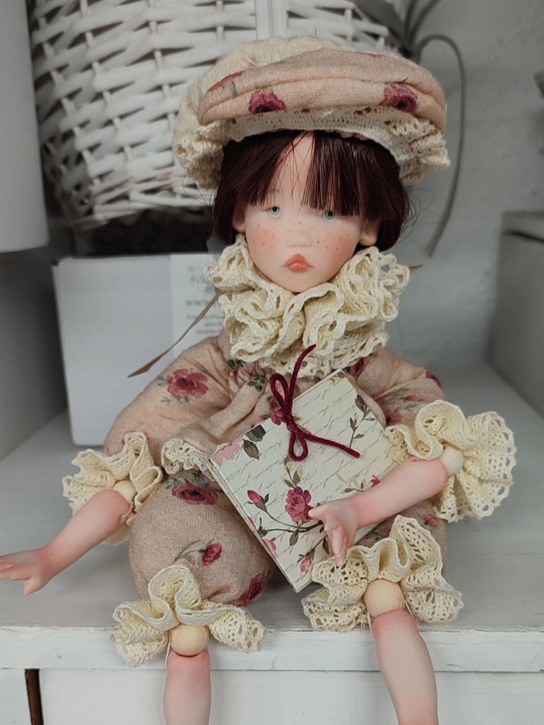 Puede incluir: Una mu&ntilde;eca hecha a mano con la cara pintada, vestida con un vestido con estampado floral, un sombrero a juego y un cuello y pu&ntilde;os con volantes. La mu&ntilde;eca sostiene un peque&ntilde;o libro con un dise&ntilde;o floral. La mu&ntilde;eca est&aacute; sentada sobre una superficie blanca.