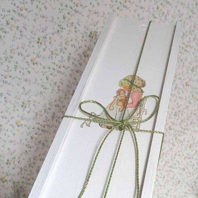 Puede incluir: Una caja de madera blanca con una ilustraci&oacute;n en acuarela de una ni&ntilde;a sosteniendo una mu&ntilde;eca, atada con hilo verde y dorado. La ilustraci&oacute;n presenta a una ni&ntilde;a con un vestido rosa y un sombrero verde. Fondo con estampado floral.