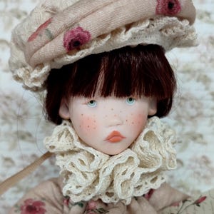 Puede incluir: Una mu&ntilde;eca con tez p&aacute;lida, pecas y ojos azules. La mu&ntilde;eca lleva un sombrero beige con un estampado floral y un cuello con volantes. La ropa de la mu&ntilde;eca tiene un dise&ntilde;o floral a juego. El pelo de la mu&ntilde;eca es casta&ntilde;o oscuro.