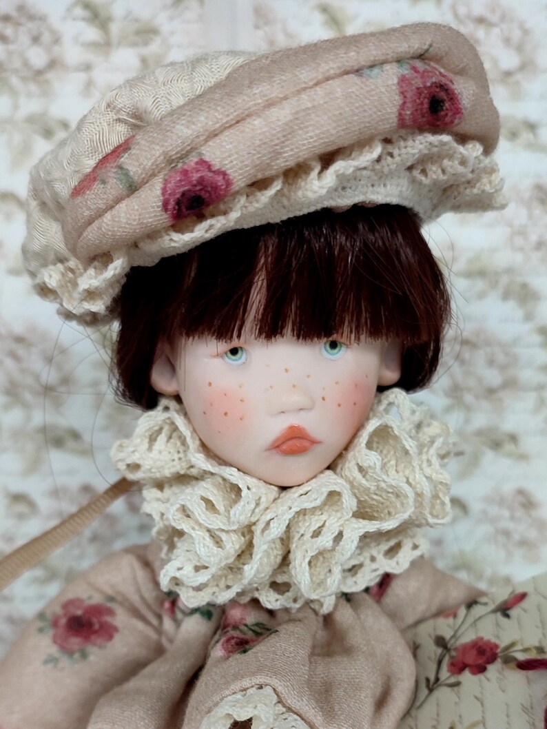 Puede incluir: Una mu&ntilde;eca con tez p&aacute;lida, pecas y ojos azules. La mu&ntilde;eca lleva un sombrero beige con un estampado floral y un cuello con volantes. La ropa de la mu&ntilde;eca tiene un dise&ntilde;o floral a juego. El pelo de la mu&ntilde;eca es casta&ntilde;o oscuro.