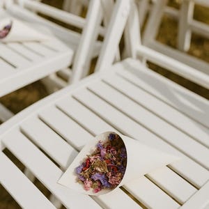 May include: White wooden folding chairs with a cone of dried flowers. The cone is filled with purple, pink, and red dried flowers. The chairs are arranged outdoors, possibly for a wedding or event.