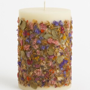 May include: A cream-colored pillar candle adorned with dried flowers. The candle is cylindrical, with a white wick at the top. The flowers are a mix of pink, purple, orange, and yellow, and are embedded around the candle's exterior.