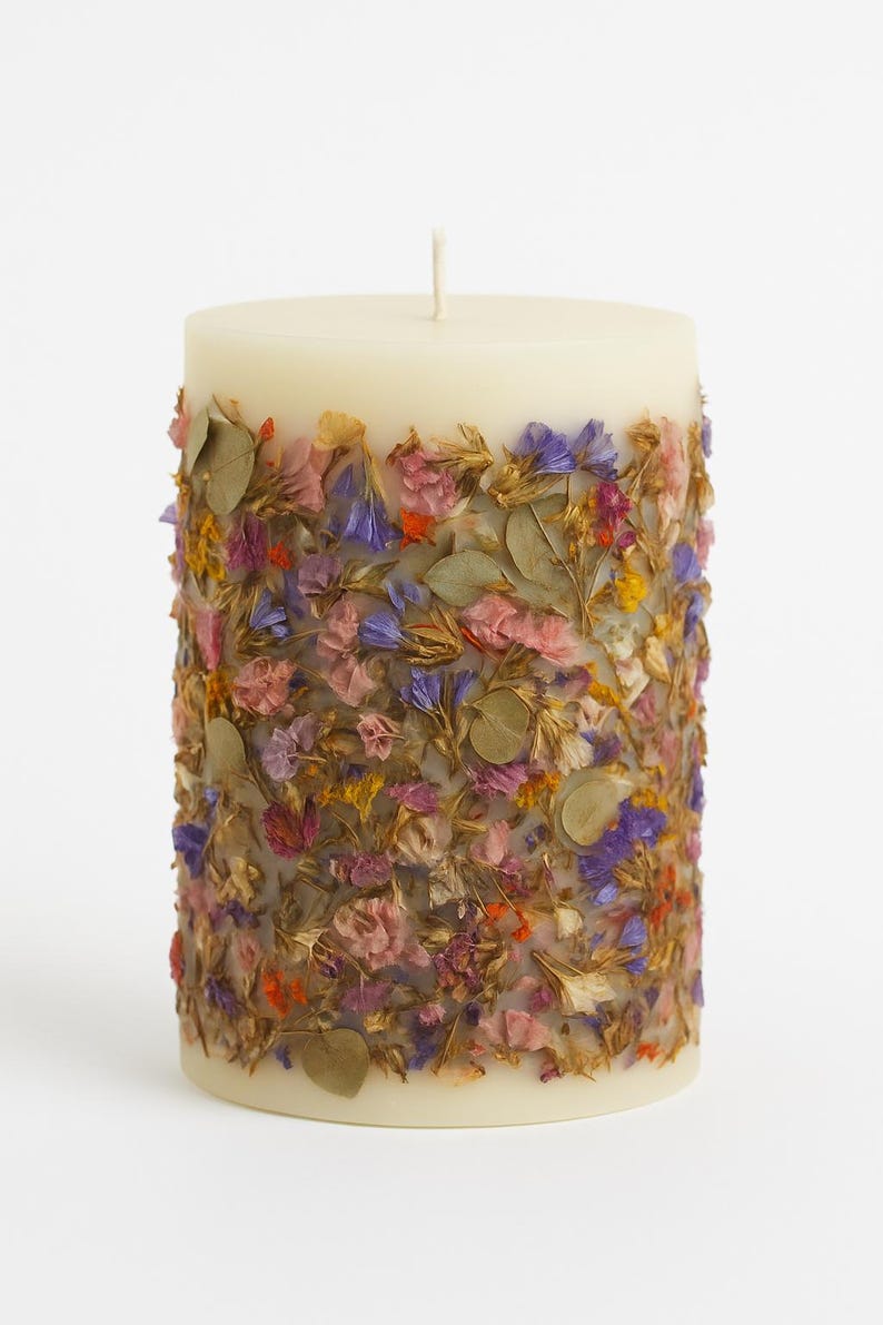 May include: A cream-colored pillar candle adorned with dried flowers. The candle is cylindrical, with a white wick at the top. The flowers are a mix of pink, purple, orange, and yellow, and are embedded around the candle's exterior.