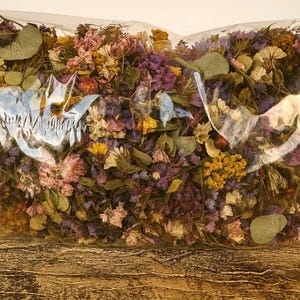 May include: A clear plastic bag filled with dried flowers in various colors, including purple, yellow, pink, and green. The flowers are tightly packed together, creating a dense and textured appearance. The bag is sitting on a wooden surface.