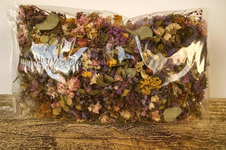 May include: A clear plastic bag filled with dried flowers in various colors, including purple, yellow, pink, and green. The flowers are tightly packed together, creating a dense and textured appearance. The bag is sitting on a wooden surface.