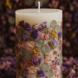 May include: A white pillar candle adorned with pressed flowers in shades of purple, pink, yellow, and green. The candle has a single white wick and is surrounded by dried flowers.