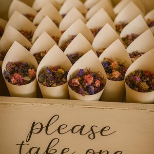 May include: A display of paper cones filled with dried flowers in shades of pink, purple, orange, and yellow. The cones are arranged in a wooden box with the words "please take one" written on the front.