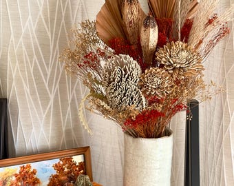 Könnte beinhalten: Eine weiße Keramikvase mit strukturierter Oberfläche beherbergt eine getrocknete Blumenarrangements. Die Anordnung umfasst getrocknete Palmblätter, getrocknete Gräser und getrocknete Blumen in Braun-, Beige- und Rottönen. Ein kleiner orangefarbener Kürbis steht vor der Vase.