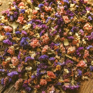 May include: A close-up of dried flowers in shades of purple, pink, and orange, scattered on a rustic wooden surface. The flowers are small and delicate, with a variety of textures and shapes. The overall effect is one of natural beauty and vintage charm.