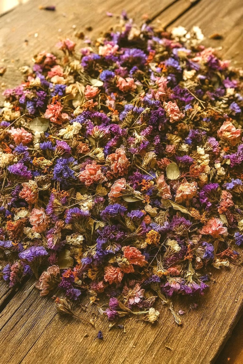 May include: A close-up of dried flowers in shades of purple, pink, and orange, scattered on a rustic wooden surface. The flowers are small and delicate, with a variety of textures and shapes. The overall effect is one of natural beauty and vintage charm.