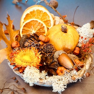 Może przedstawiać: Rustykalny kosz przepełniony dekoracjami w jesiennym klimacie. Zawiera małą dynię, suszone plasterki pomarańczy, szyszki, żołędzie i jesienne liście w odcieniach pomarańczu, żółci i brązu. Aranżację uzupełnia biały mech.