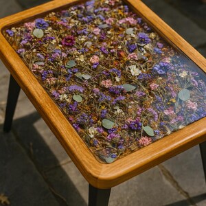 May include: A rectangular coffee table with a clear resin top filled with dried flowers in shades of purple, pink, and white. The table has a light brown wooden frame and black legs.