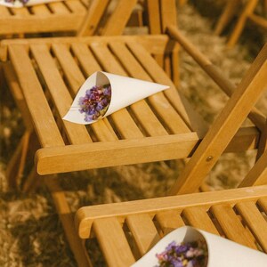 May include: Wooden folding chairs with white paper cones filled with dried purple and pink flowers. The chairs are arranged outdoors, likely for a wedding or special event. The natural wood has a warm tone.
