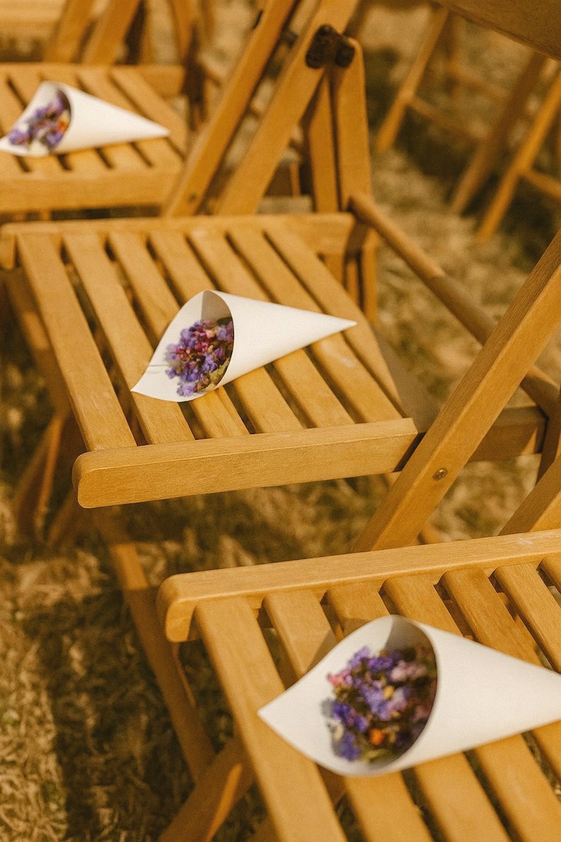 May include: Wooden folding chairs with white paper cones filled with dried purple and pink flowers. The chairs are arranged outdoors, likely for a wedding or special event. The natural wood has a warm tone.