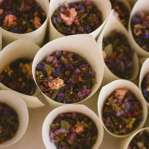 May include: Close-up of several white paper cones filled with dried flowers. The flowers are a mix of purple, pink, and yellow hues. The cones are arranged on a white surface, creating a visually appealing display, perfect for weddings.