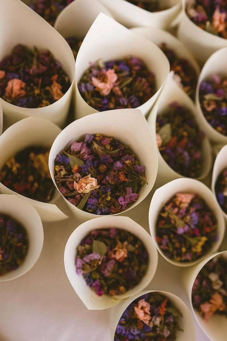 May include: Close-up of several white paper cones filled with dried flowers. The flowers are a mix of purple, pink, and yellow hues. The cones are arranged on a white surface, creating a visually appealing display, perfect for weddings.