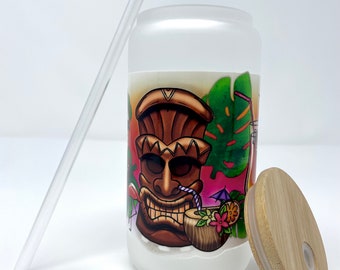 May include: A frosted glass tumbler with a tiki-themed design featuring a brown tiki face, palm leaves, and a coconut with a straw. The tumbler has a bamboo lid and comes with a clear plastic straw.