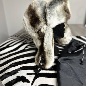 May include: A faux fur trapper hat in shades of brown, grey, and white, with ear flaps and a chin strap. The hat is displayed on a black mannequin head. A black drawstring bag is next to the hat.