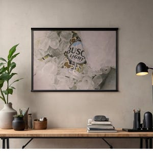 May include: A framed print featuring a bottle of Busch Light beer submerged in ice. The print is displayed above a wooden desk with a lamp, books, and office supplies. A green plant in a vase is on the left.