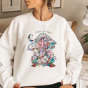 May include: White sweatshirt featuring a colorful illustration of mushrooms, flowers, and crystals. The design includes the text "Made of magic" in cursive script. The artwork has watercolor-style splashes of color.